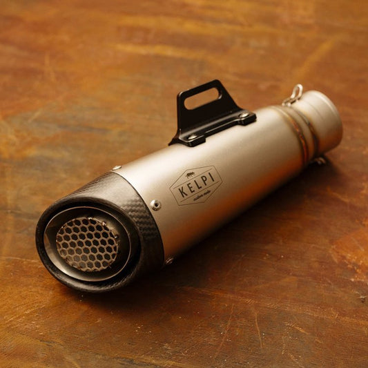 A carbon-fiber Kelpi Flanker muffler with a stainless steel shell, carbon fibre end cap, and a black mounting bracket on a wooden surface.