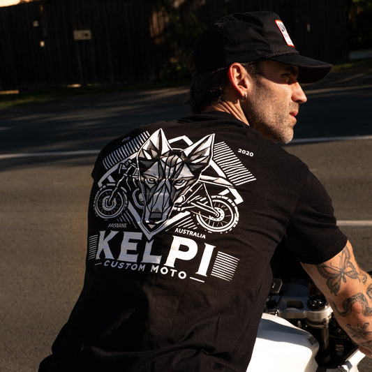 Man sitting on a white motorcycle wearing a black Kelpi Custom Moto t-shirt.
