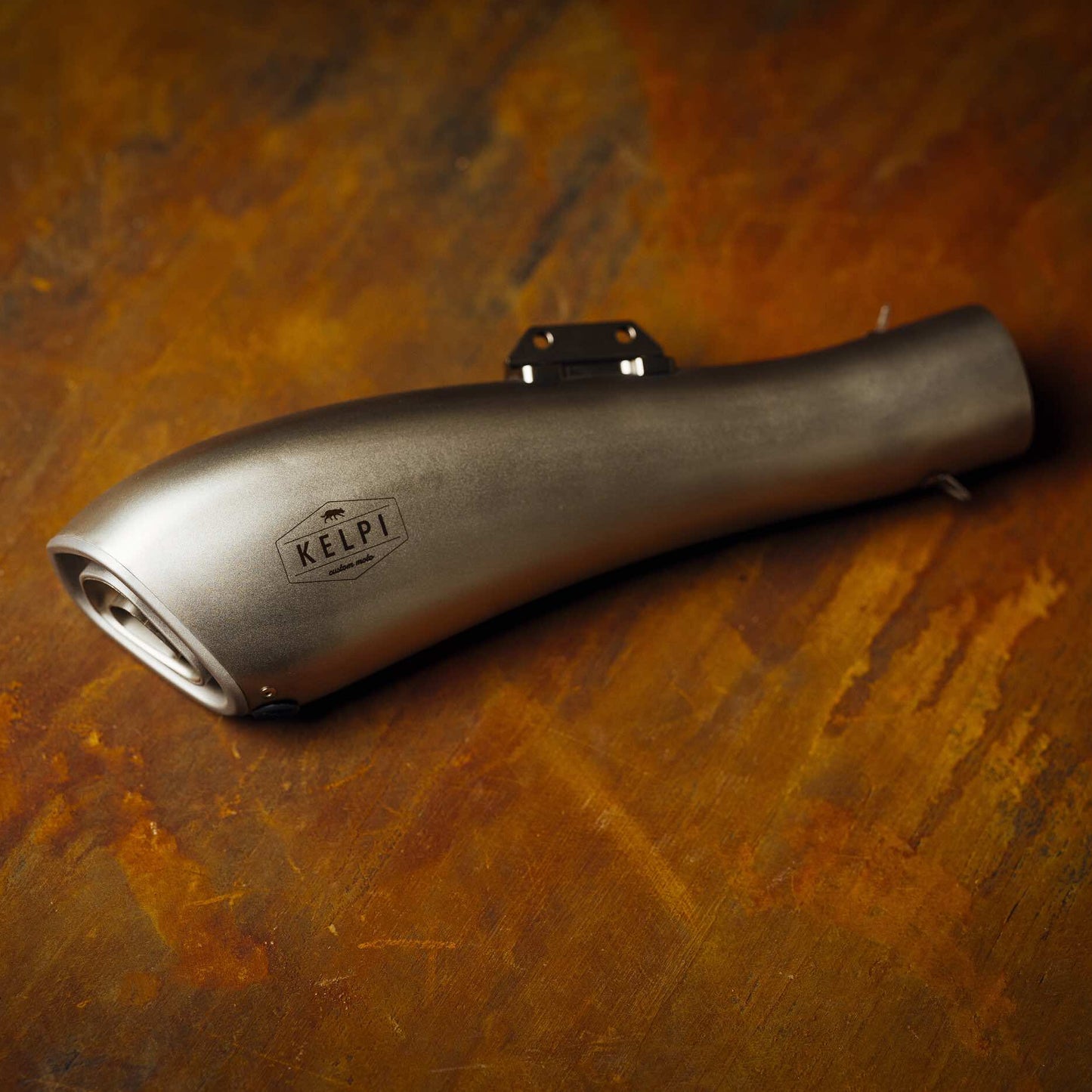 A stainless steel Alpha Muffler with a hydroformed shape, placed on a textured brown surface.