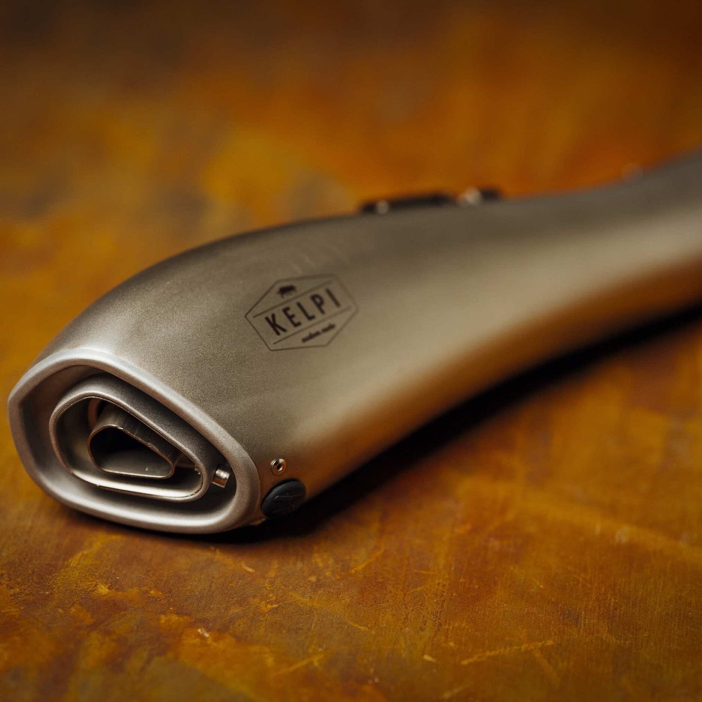 A stainless steel Alpha Muffler with a hydroformed shape, placed on a textured brown surface.