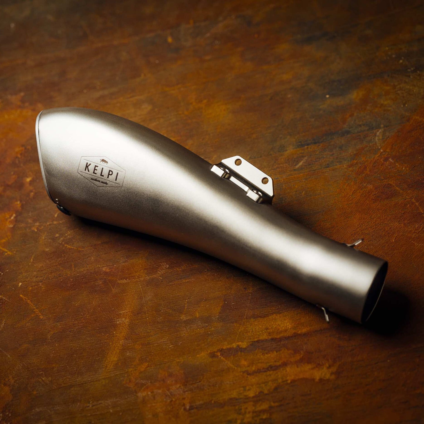 A stainless steel Alpha Muffler with a hydroformed shape, placed on a textured brown surface.
