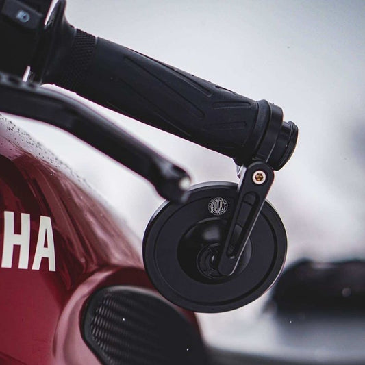 Close-up image of a Hindisght bar end motorcycle mirror attached to a motorcycle handlebar, with a blurred red motorcycle body in the background.