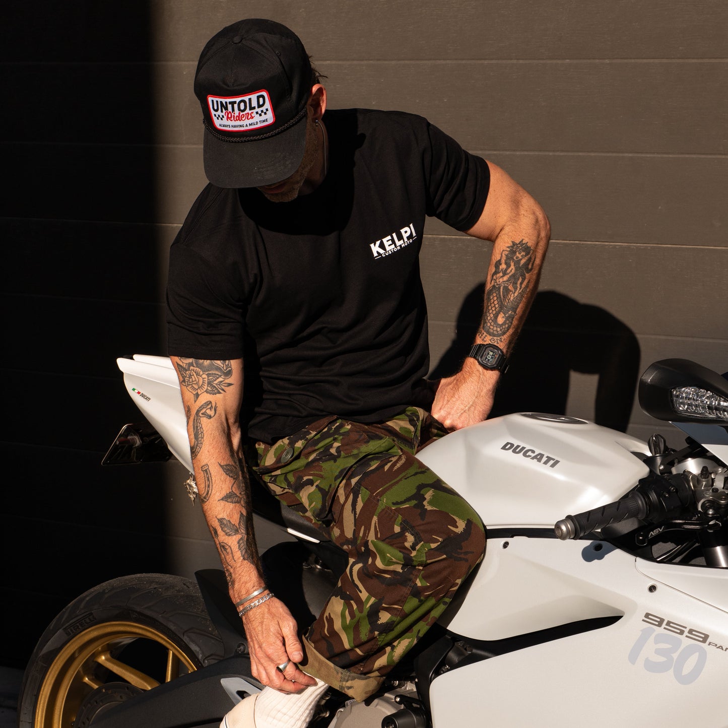 Man sitting on a white Ducati motorcycle wearing a black t-shirt and camouflage pants.