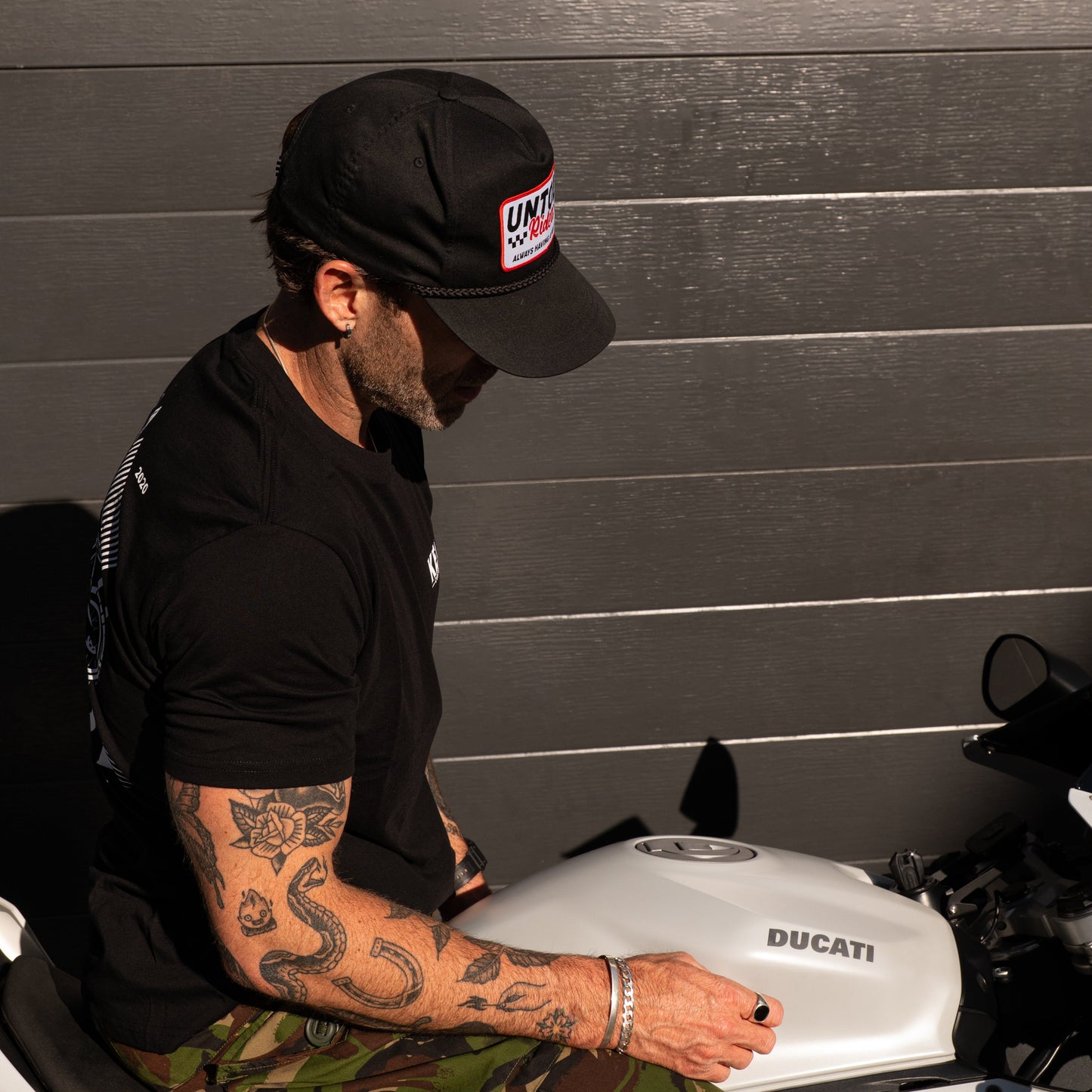 Person wearing a black cap and black shirt sitting on a Ducati motorcycle in a garage.